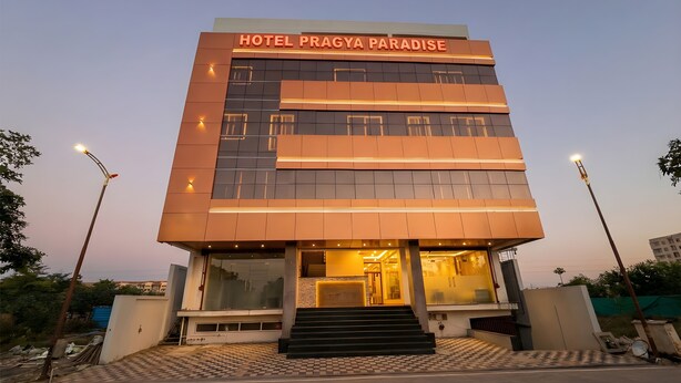 Edifici modern d'hotel anomenat "HOTEL PRAGYA PARADISE" amb façana de vidre, una entrada ben il·luminada i una escala que condueix al vestíbul.
