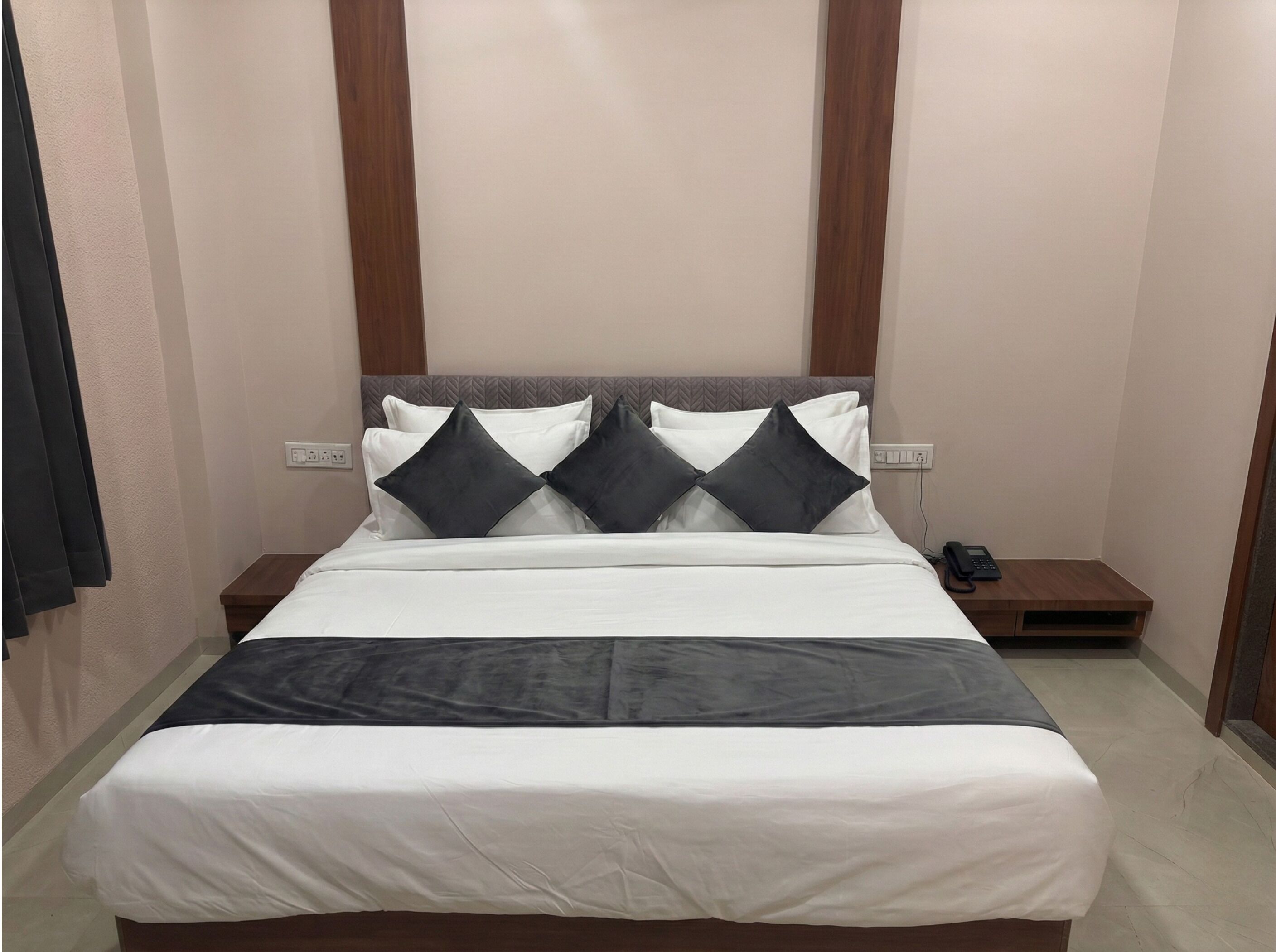 Una cama bien hecha con colcha blanca y gris, dos almohadas negras y un cabecero de madera. Se observa un mesita de noche con un teléfono y un enchufe en la pared junto a la cama.