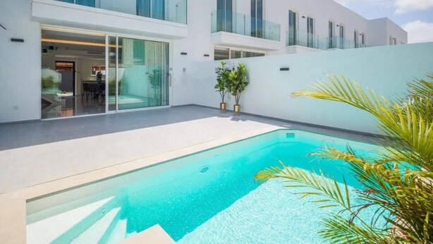 A modern outdoor swimming pool with a clear blue water, surrounded by a white building with large windows and a glass door entrance. - Alma Nomada Luxury Villa & Private Pool & BBQ & Solarium (Corralejo)
