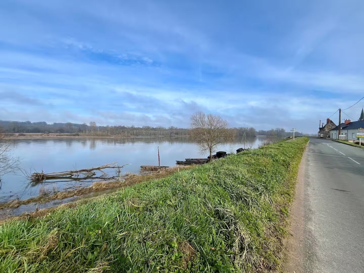 Une route de campagne longe un lac paisible, avec une berge herbeuse et un petit bâtiment visible au loin.