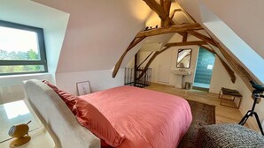Et soveværelse med skrå vægge, en seng med pink dyne, et skrivebord med lampe, et spejl og et vindue med udsigt til træer.
