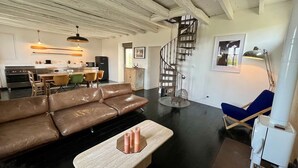 En rummelig opholdsstue med en brun lædersofa, et træbord og en blå stol. En spiraltrappe fører op til en etage over.