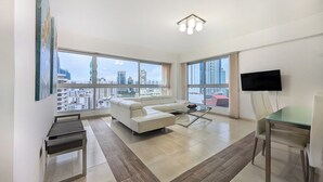 A modern living room with a white sofa, a glass coffee table, and a TV mounted on the wall. Large windows offer a city view. - YC 1701l Apartamento 2 BDR Av Balboa (Panama City)