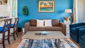 A living room with a blue wall, a brown sofa, a coffee table, and a blue armchair. - Island Surf 304 - Modern Renovation, Must See Ocean View, Cool Split AC, Wi-fi ! (Kihei)