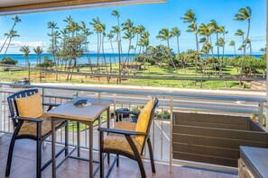 A balcony with a view of the ocean and palm trees, a table with chairs, and a railing. - Island Surf 304 - Modern Renovation, Must See Ocean View, Cool Split AC, Wi-fi ! (Kihei)