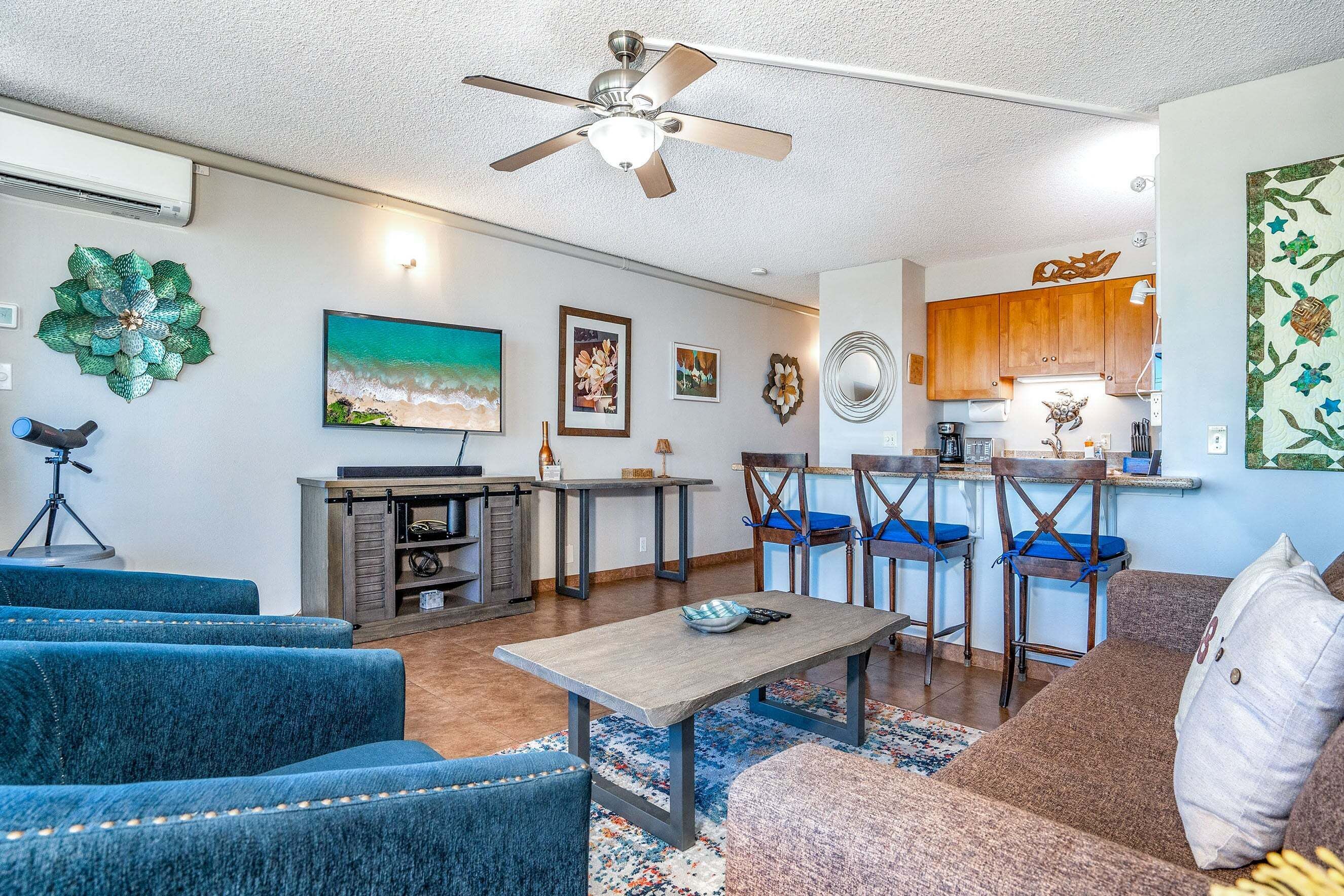 A modern living room with a sofa, a coffee table, and a TV. There is a kitchen area with wooden cabinets and a bar stool.