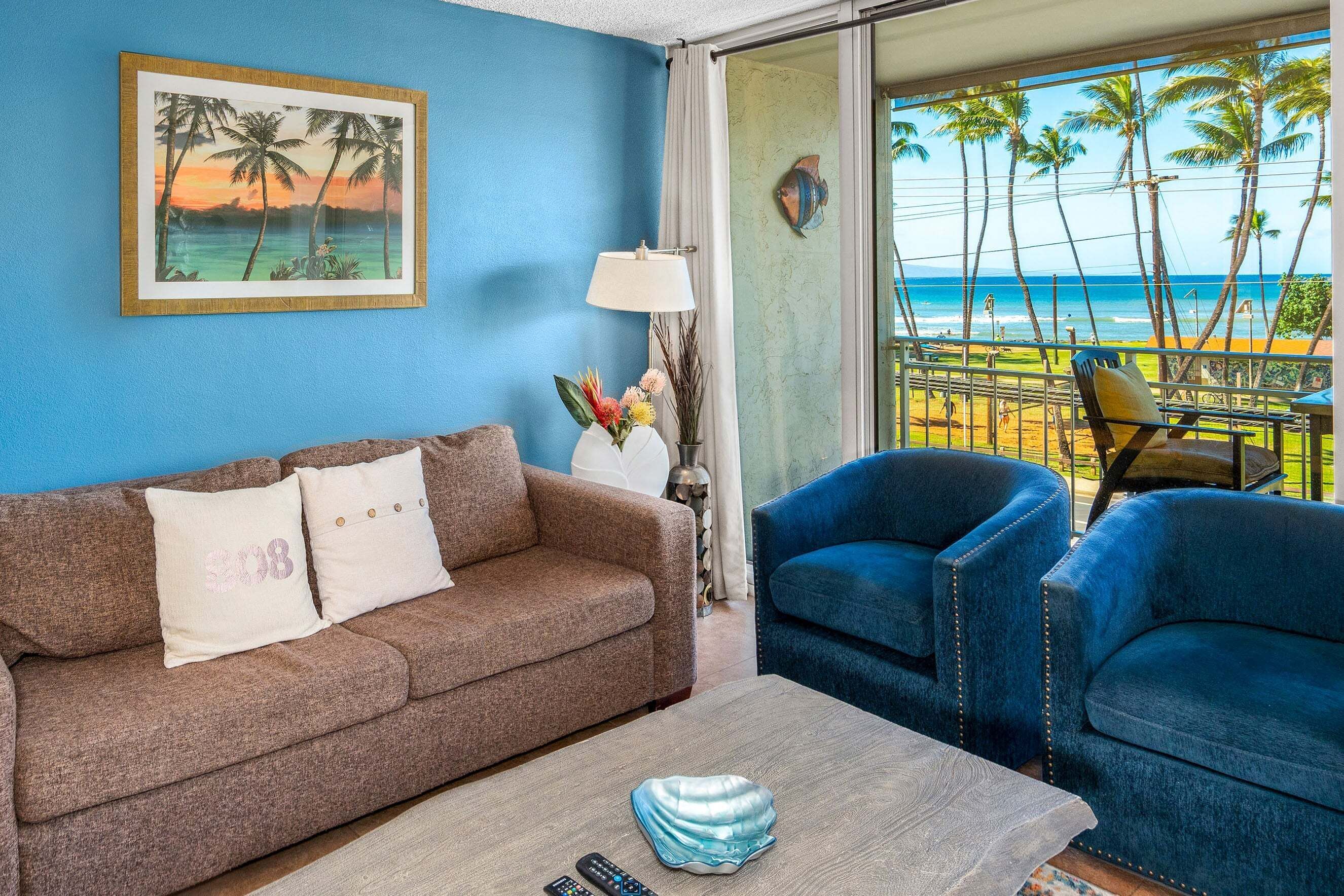 Ein Wohnzimmer mit einem braunen Sofa, zwei blauen Sesseln, einem Couchtisch mit einer Muschel und einem Gemälde, das einen Sonnenuntergang über dem Meer zeigt.