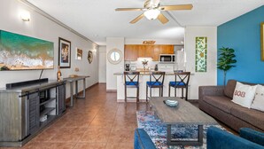 A modern living room with a sofa, coffee table, and TV. There is a kitchen area with wooden cabinets and a microwave. The room has a ceiling fan and a large painting on the wall. - Island Surf 304 - Modern Renovation, Must See Ocean View, Cool Split AC, Wi-fi ! (Kihei)