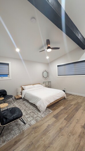 A bedroom with a bed, a ceiling fan, a round mirror, a small table, a chair, and a window with blinds.