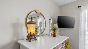 Un tocador blanco con espejo, una vela encendida y objetos decorativos. Un televisor de pantalla plana está montado en la pared.