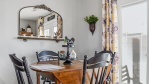 Un comedor con mesa de madera, sillas negras, un espejo, una estantería con objetos decorativos y una ventana con cortinas.