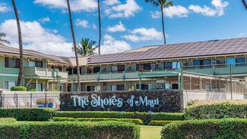 A hotel with a sign reading 'The Shores of Maui,' featuring a multi-story building with balconies, a stone wall with the hotel's name, and a well-maintained garden.