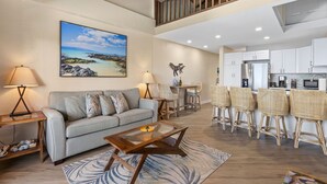 A spacious living area with a sofa, coffee table, and dining chairs. A kitchen is visible in the background with white cabinets and a dining table. - Shores of Maui 215 - Dramatic Ocean Views, Steps to the Beach, Renovated, AC! (Kihei)