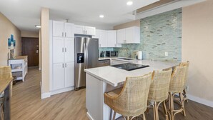 A modern kitchen with white cabinets, stainless steel appliances, and a central island with wicker chairs. - Shores of Maui 215 - Dramatic Ocean Views, Steps to the Beach, Renovated, AC! (Kihei)