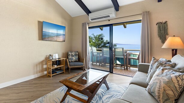 A living room with a glass coffee table, a sofa, a chair, a small wooden side table, and a painting on the wall. - Shores of Maui 215 - Dramatic Ocean Views, Steps to the Beach, Renovated, AC! (Kihei)