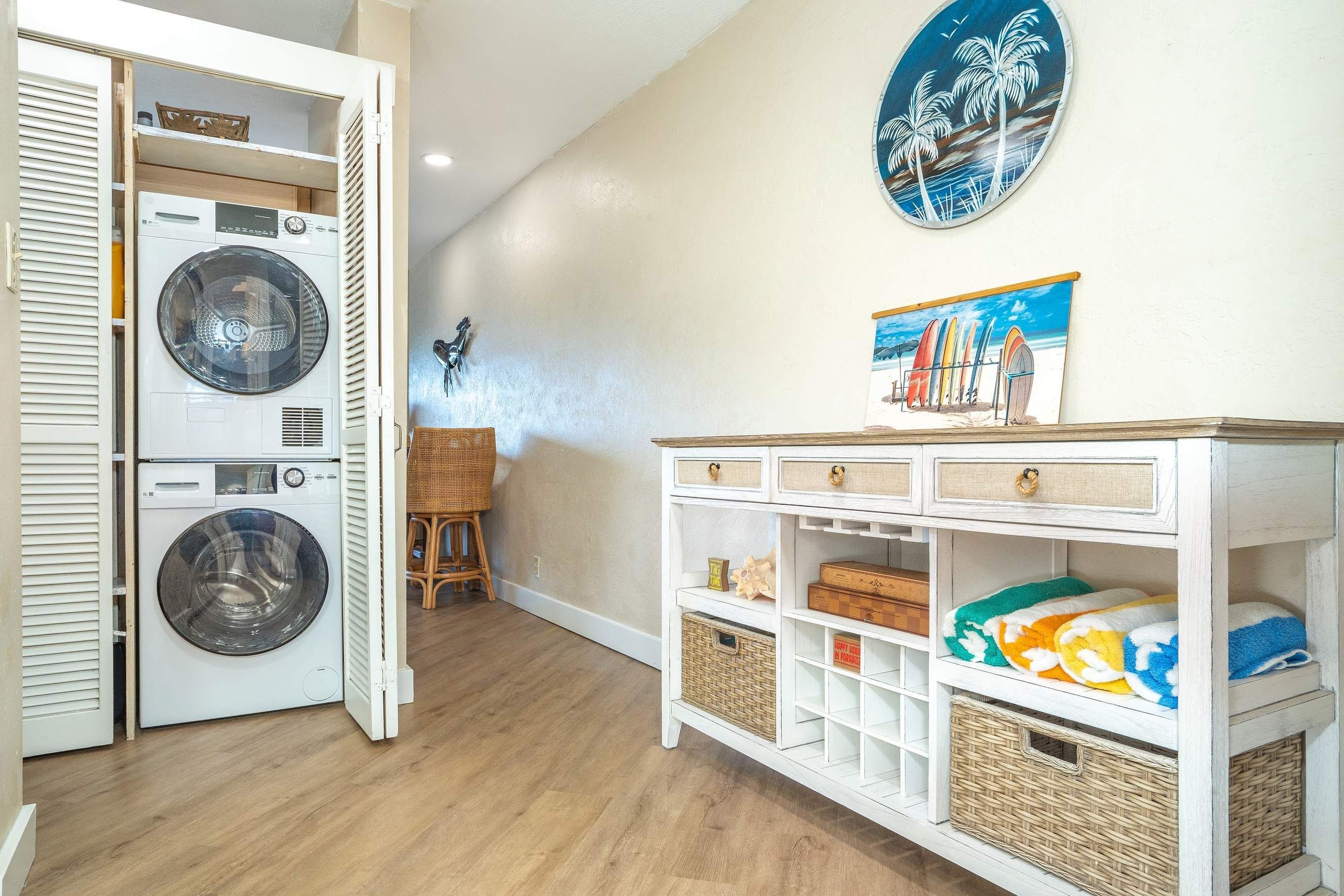 A laundry room with a washing machine and dryer, a wicker chair, and a wall-mounted picture of palm trees.