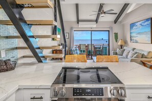 A modern kitchen with a central island, a dining area with wicker furniture, and a large window overlooking the sea. - Shores of Maui 215 - Dramatic Ocean Views, Steps to the Beach, Renovated, AC! (Kihei)