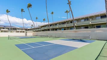 A tennis court with a net, surrounded by palm trees and a building with multiple balconies.
