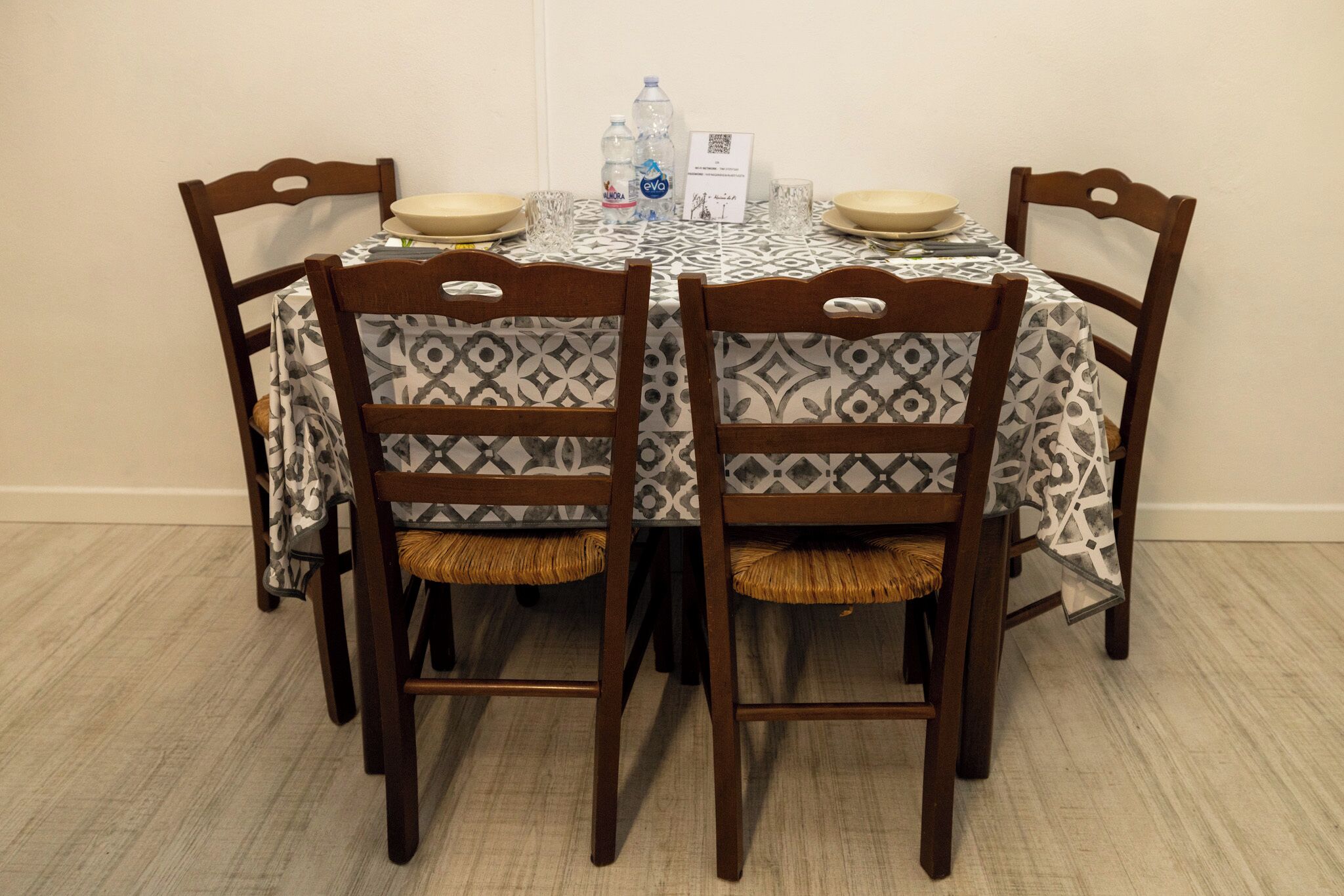 Une table à manger recouverte d’une nappe à motifs, entourée de chaises en bois dont les assises sont garnies de coussins tissés.