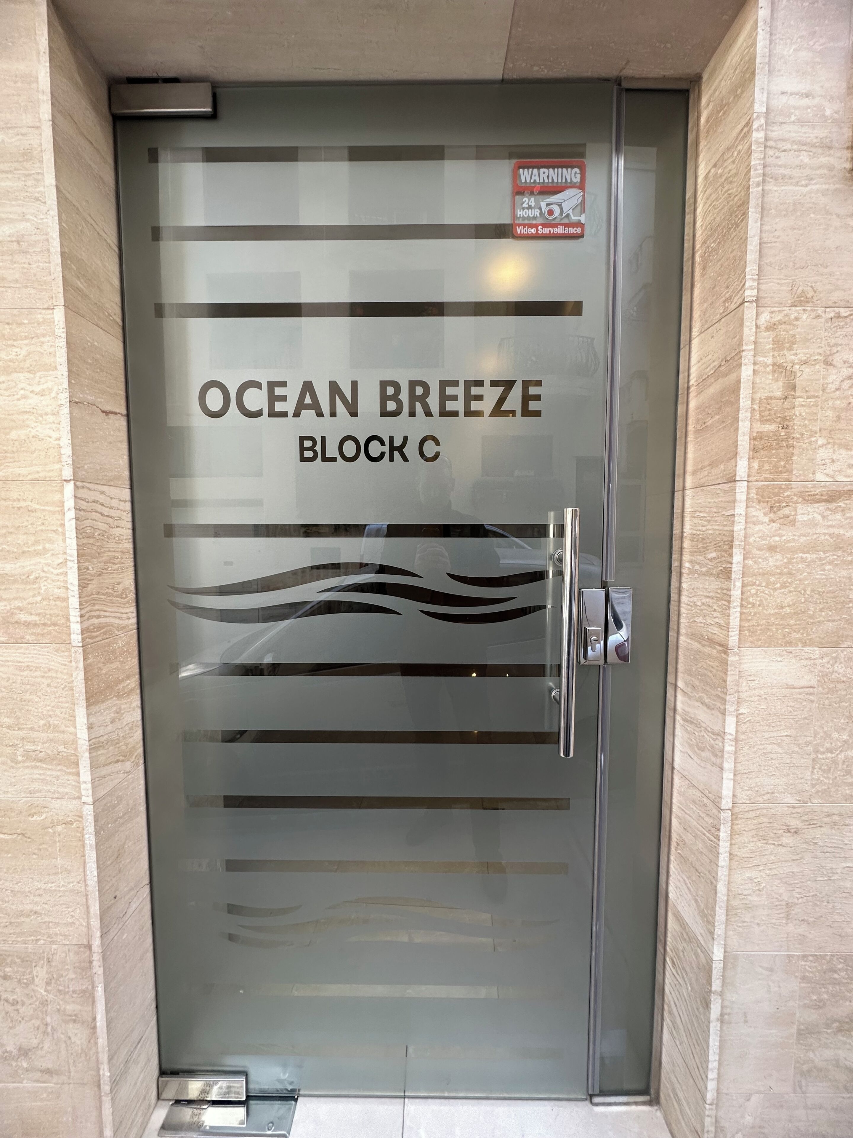 A glass door with 'OCEAN BREEZE BLOCK C' written on it, a warning sign for 24-hour video surveillance, and a metal handle.