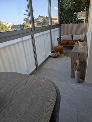 Un balcón moderno con vistas a edificios, una mesa de madera, un sofá con cojines y una planta pequeña en un atril de madera.