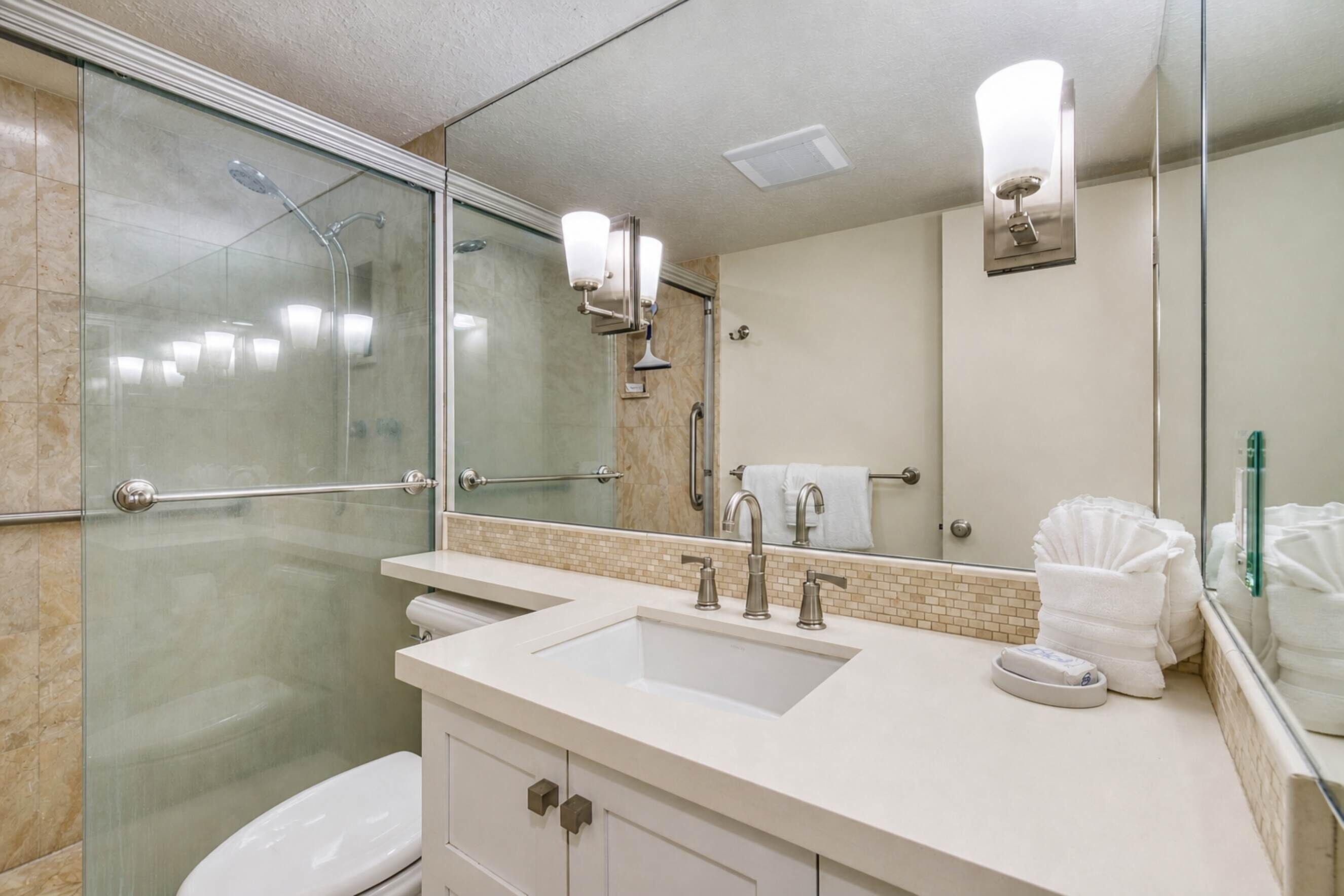 A modern bathroom with a glass shower enclosure, a white sink with two faucets, and a large mirror above the sink.