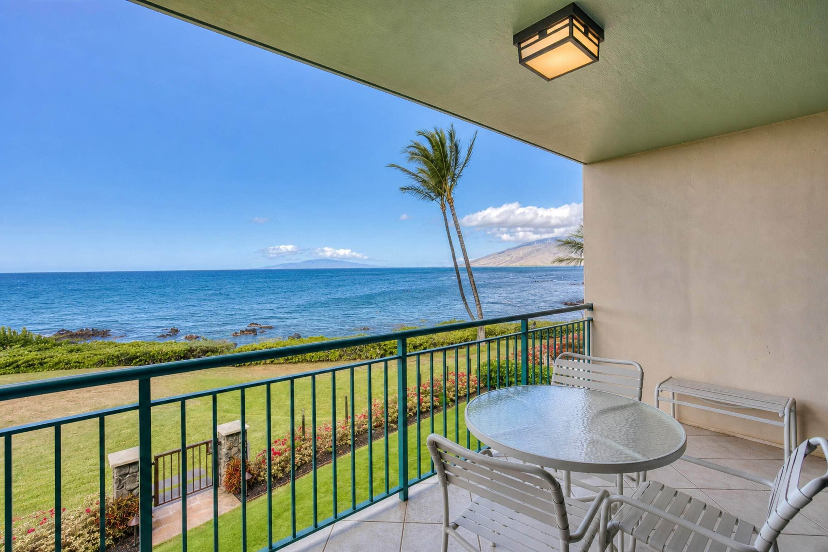 A balcony with a glass table and chairs, overlooking a beach with the ocean and a mountain in the distance.