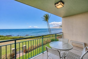 A balcony with a glass table and chairs, overlooking a beach with the ocean and a mountain in the distance. - Punahoa Beach 201 - Direct Ocean Front, Beautiful Renovation, Split AC, Wi-fi (Kihei)