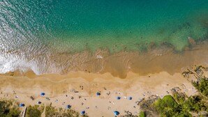 Vista aérea de una playa con aguas turquesas cristalinas, orilla arenosa y sombrillas dispersas.
