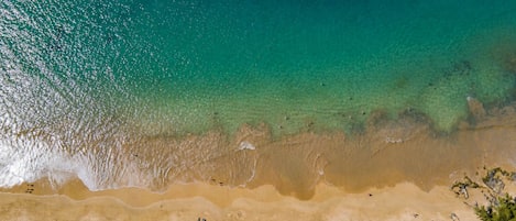 An aerial view of a beach with clear turquoise water, sandy shore, and scattered umbrellas.