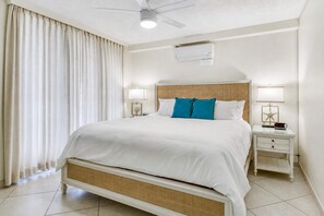 A hotel room with a bed, two bedside tables, a ceiling fan, an air conditioning unit, and a window with curtains. - Punahoa Beach 201 - Direct Ocean Front, Beautiful Renovation, Split AC, Wi-fi (Kihei)