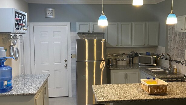 Cucina con isola centrale, frigorifero in acciaio inox, mobili bianchi e illuminazione a sospensione.