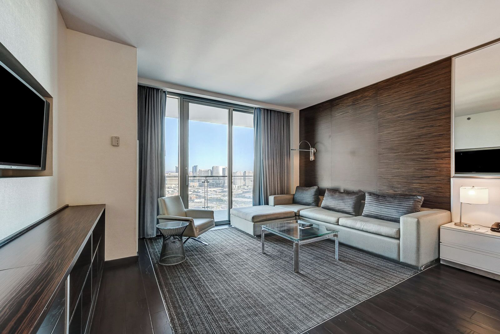 Una habitación de hotel moderna con un ventanal grande, un sofá, una silla, una mesa de centro y un televisor.