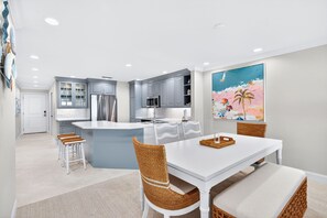 A modern kitchen with grey cabinets, a white island, and a dining area with wicker chairs and a white table. - Sanddollar B302: New Top-Floor 3BR Luxury Beachfront Condo on Sanibel (Sanibel)