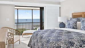 Quarto com uma cama grande, mesa de cabeceira, uma cadeira e vista para o mar através de uma porta deslizante.