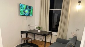 Un salon moderne avec un canapé gris, une table basse ronde noire, une petite table noire accompagnée de deux chaises, un téléviseur fixé au mur et une fenêtre avec des rideaux.