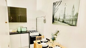 Une cuisine compacte avec des meubles blancs, une cuisinière à gaz et un dosseret carrelé. Une table à manger avec des chaises est installée dans l’espace repas adjacent, et un tableau représentant un paysage urbain est accroché au mur.
