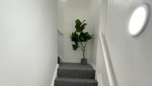 Un escalier dont les marches sont recouvertes d’une moquette grise, avec des murs blancs et un pot de fleurs placé en haut.