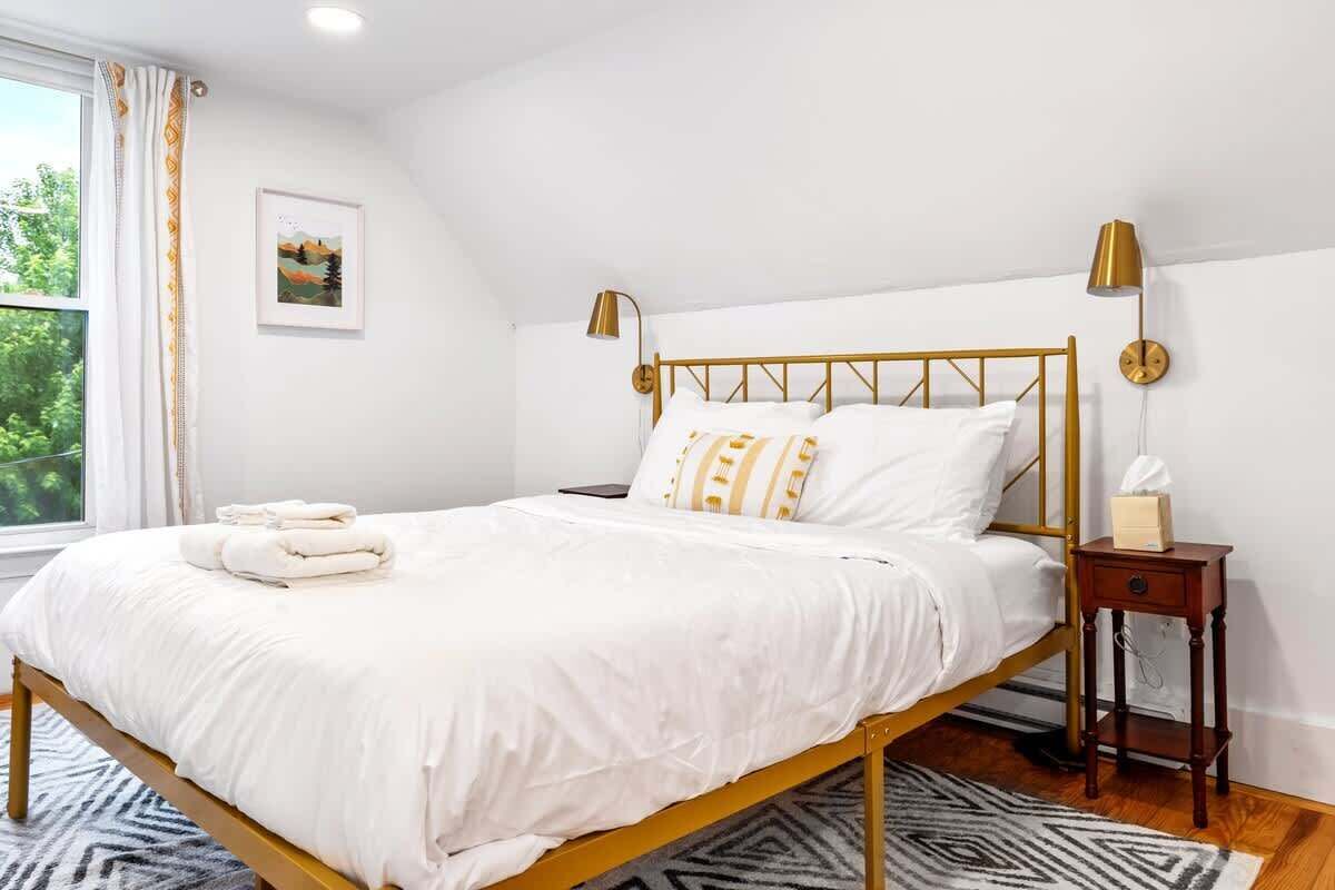 Ein ordentlich bezogenes Bett mit weißer Bettwäsche und goldenen Akzenten, ein Nachttisch mit Lampe sowie ein Fenster mit Vorhängen.