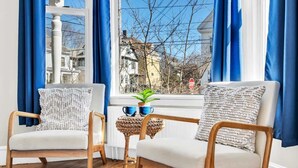 Una habitación acogedora con dos sillas de madera, una mesita de noche tejida y una ventana con cortinas azules.