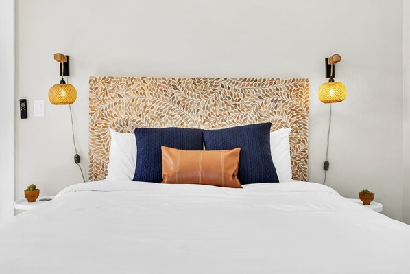 Una cama bien hecha con un cabecero estampado, dos lámparas fijadas a la pared y pequeñas macetas con plantas en las mesitas de noche.