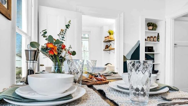 Comedor con una mesa dispuesta para dos, con un tazón blanco, vaso transparente y un jarrón con flores.