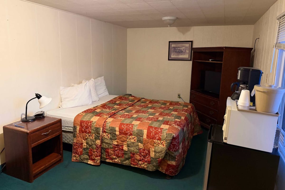 A hotel room with a bed, a nightstand, a desk with a lamp, a coffee maker, and a refrigerator.