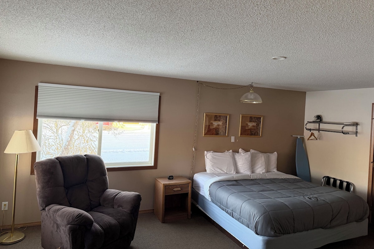 A bedroom with a bed, a chair, a nightstand, a lamp, and a window with blinds.