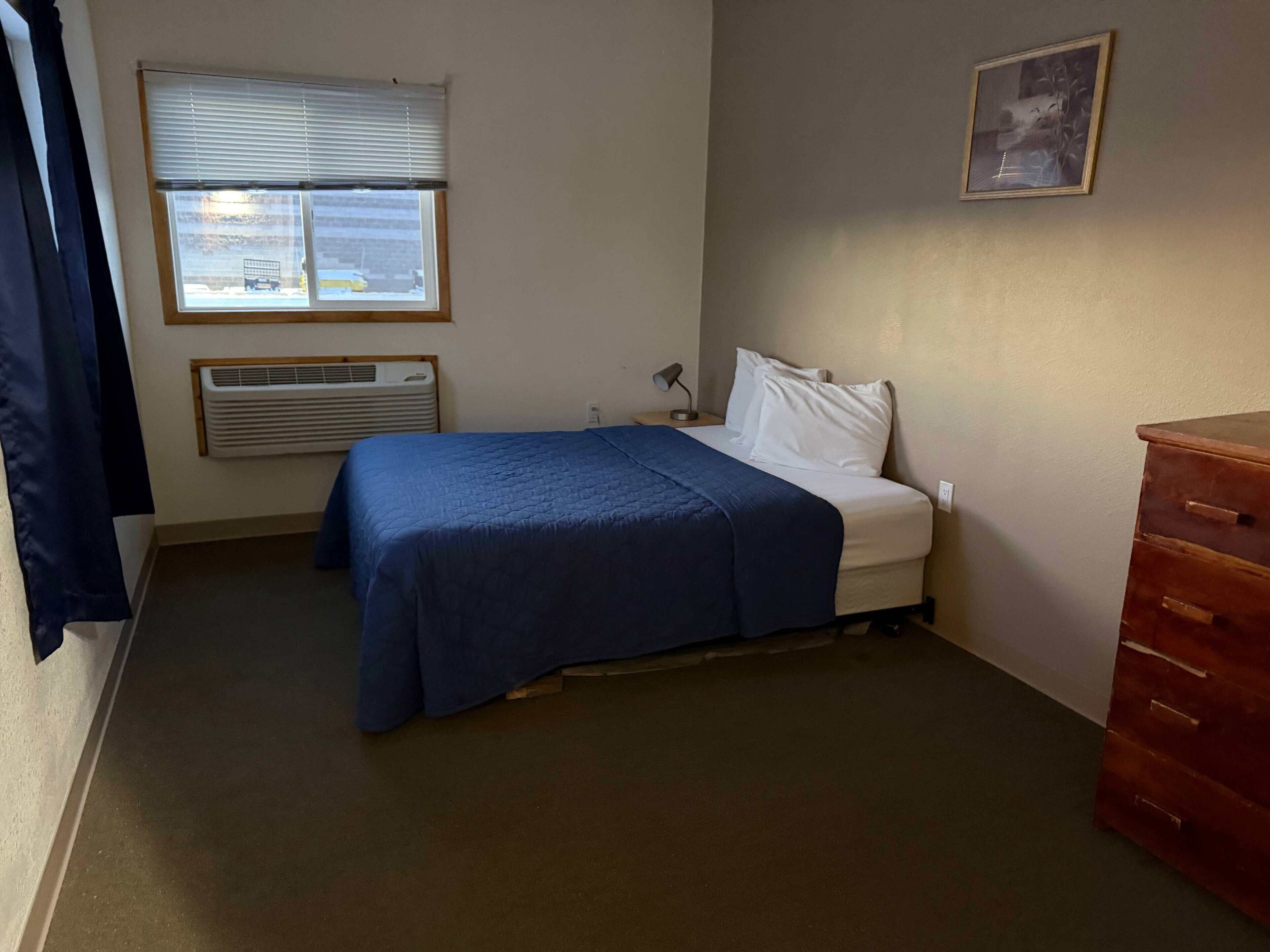 Ein Schlafzimmer mit einem Bett, einem Schminktisch, einem Fenster mit Jalousien, einer Klimaanlage und einem Bild an der Wand.