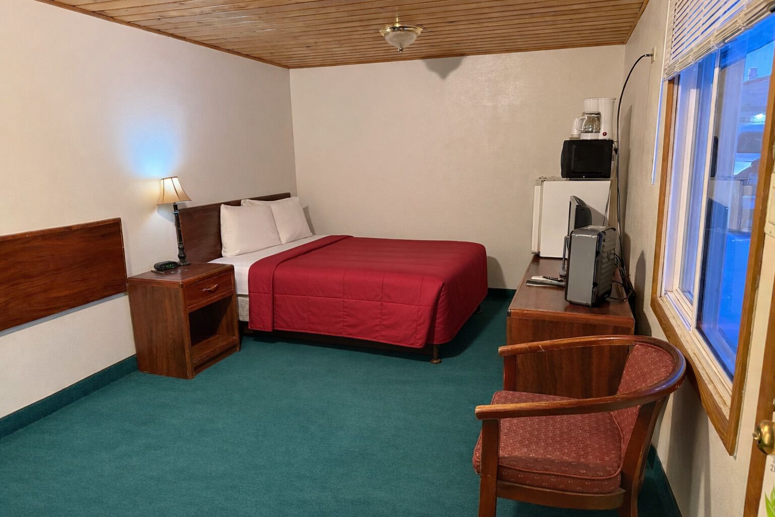 A hotel room with a bed, a desk with a computer, a chair, a nightstand, and a window with blinds.