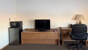 A compact hotel room with a wooden desk, a television, a small refrigerator, a lamp, and a black office chair. - Sagebrush Inn (Baker)