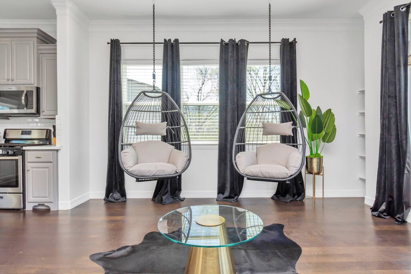 A modern living room with two hanging chairs, a glass coffee table, and a kitchen in the background.