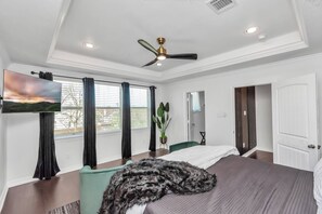 Un dormitorio con cama, ventilador de techo, televisor montado en la pared y ventana con cortinas.