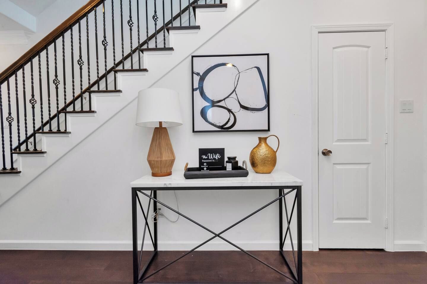 A staircase with black balusters, a white door, a table with a lamp, a vase, and a framed abstract painting.